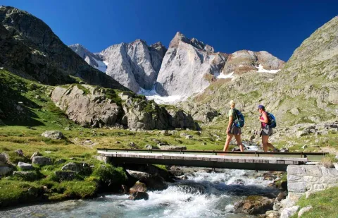 Deux randonneurs franchissent une passerelle face au massif du Vignemale dans les Pyrénées
