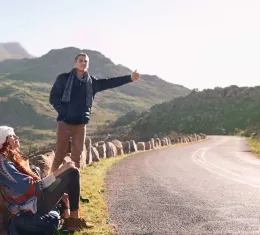 Deux voyageurs font de l'auto-stop au bord d'une route de montagne en attendant qu'un automobiliste s'arrête.