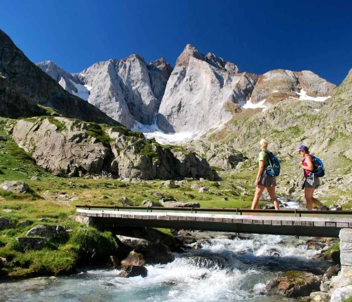 Deux randonneurs franchissent une passerelle face au massif du Vignemale dans les Pyrénées