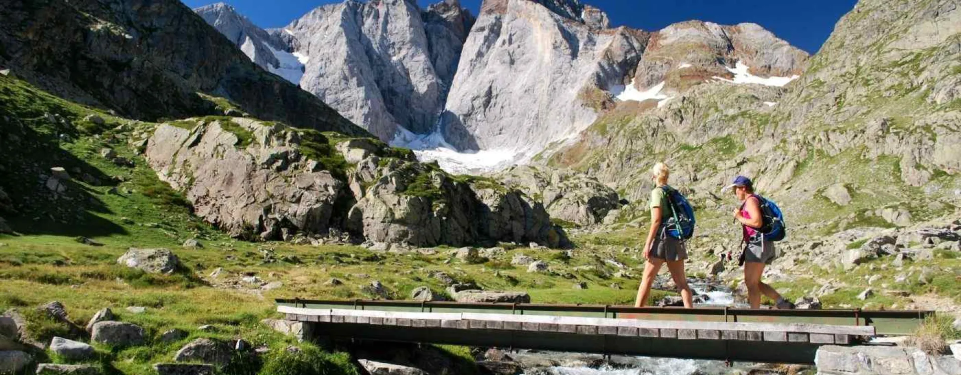 Deux randonneurs franchissent une passerelle face au massif du Vignemale dans les Pyrénées