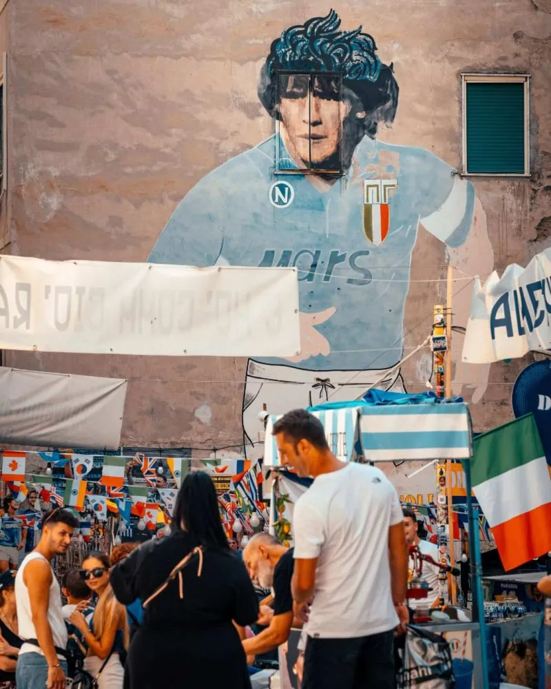 Fresque de Diego Maradona dans une rue animée de Naples avec des habitants et des drapeaux