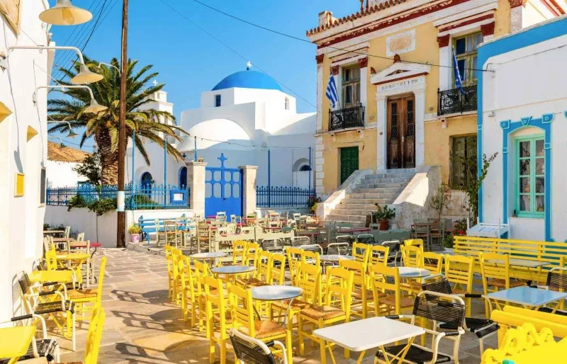Place calme à Hora sur l’île de Serifos avec tables et chaises colorées devant une église blanche typique des Cyclades