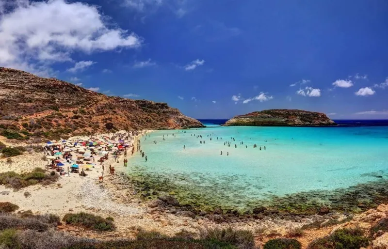 Baigneurs sur la Spiaggia dei Conigli à Lampedusa avec ses eaux turquoise et l’îlot des Conigli en arrière-plan