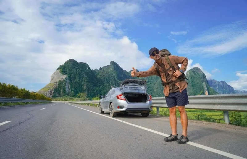 Auto-stoppeur au bord d'une route à côté d'un véhicule en panne, illustrant les risques et dangers de l'auto-stop lors d'un voyage.
